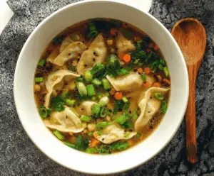 A steaming bowl of Asian Dumpling Potsticker Soup featuring golden potstickers, fresh green vegetables, and flavorful broth garnished with chopped scallions.